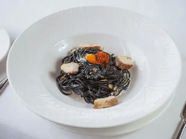 Pasta negra con trozos de pescado, huevas de salmón y yema de huevo. La tinta de calamar, ingrediente principal, aporta un sabor único y propiedades beneficiosas.