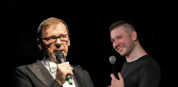 Dwóch mężczyzn z mikrofonami na scenie. Jeden w garniturze, drugi w czarnej koszulce. Widać, że socha stand up.