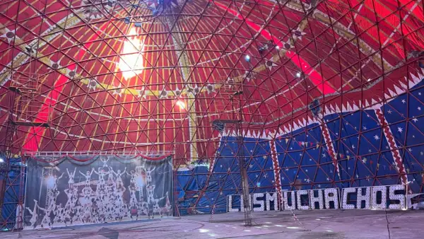 Interior de carpa de circo con techo rojo y azul, telón de fondo de acróbatas y luces que forman "MUCHACHOS". ¡Un espectáculo en Bempota, Ourense!
