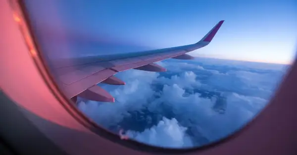 View from an airplane window at sunset. The wing stretches across the frame, bathed in pink and purple light, over a sea of clouds.