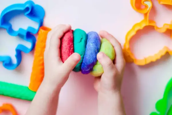 Kinderhände halten bunte Knetmasse, bereit zum Kneten. Nebenan liegen bunte Ausstecher, perfekt zum Knetseife selber machen.