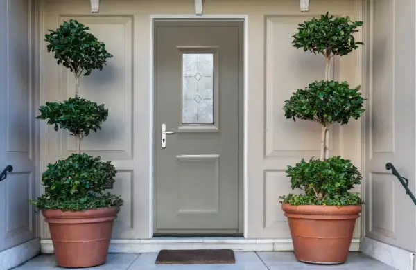 Une porte d'entrée grise avec une fenêtre décorative, encadrée par deux grands pots de plantes taillées. Idéal pour savoir comment changer une porte d'entrée.