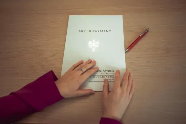 Dłonie na akcie notarialnym, obok czerwony długopis. Dokument zawiera pieczęć państwową i dane notariusza.
