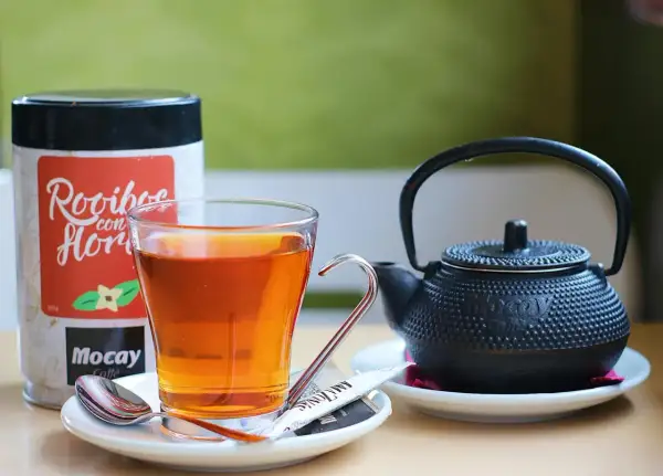 Szklanka z bursztynowym naparem, obok czajniczek i puszka z napisem "Rooibos con Flor". Czas na chwilę relaksu z rooibos.