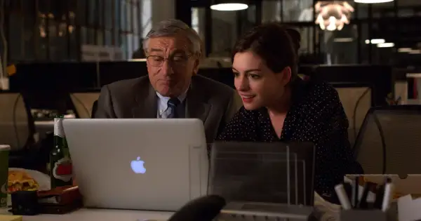 Robert De Niro i Anne Hathaway pracują przy laptopie. Młoda praktykantka z entuzjazmem analizuje dane, podczas gdy doświadczony szef czuwa nad jej pracą.