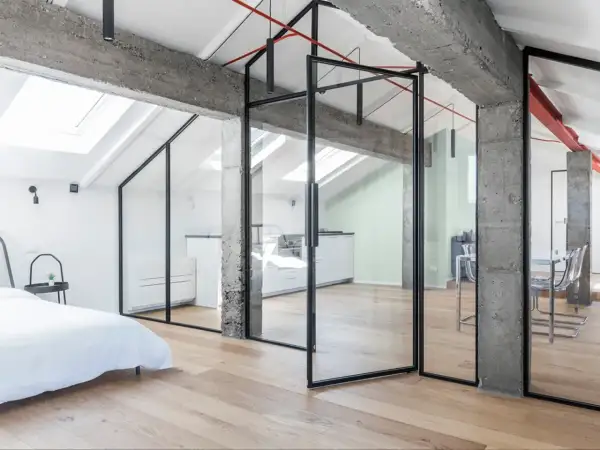 Moderno recupero sottotetto con pareti in vetro, travi a vista in cemento e parquet chiaro. Ambiente luminoso con cucina e zona living.