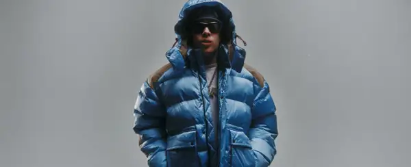 Giovane con piumino blu acceso, cappuccio alzato, occhiali da sole e una collana con ciondolo. Lo sfondo è un grigio chiaro, quasi low red.