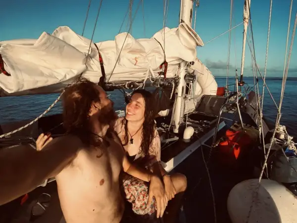 Coppia felice in barca a vela al tramonto, con vele bianche spiegate e mare calmo.