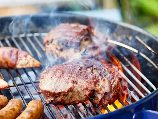 Saftiges Fleisch zum Grillen zischt auf dem Rost. Würstchen und Steaks brutzeln über heißen Kohlen, umgeben von Rauch und Flammen.