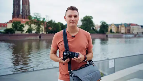 Mężczyzna z aparatem w rękach, w plenerze nad rzeką, z widokiem na zabytkowe budynki i katedrę.
