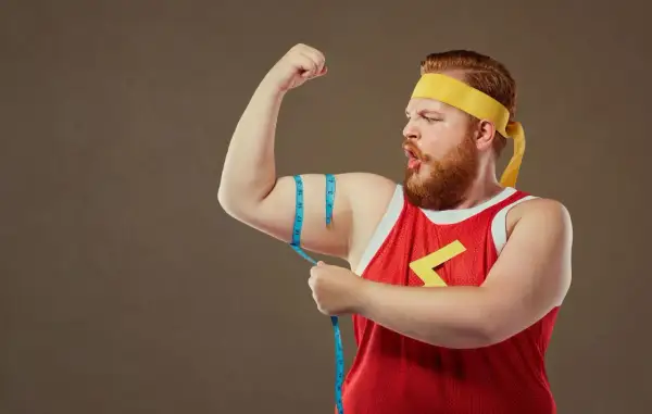 Mężczyzna typu endomorfik z wąsami i brodą, w żółtej opasce i czerwonej koszulce, mierzy obwód bicepsa.