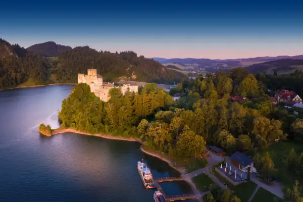 Zamek na skale nad jeziorem, otoczony lasem. Widać łodzie przy pomoście. To jedne z fajnych miejsc nad jeziorem.