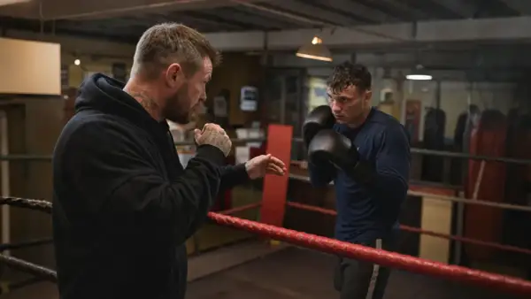 A boxer trains with his coach in a boxing ring. The coach, with tattoos on his arms and neck, demonstrates a move. The boxer, wearing gloves, listens intently. This is a scene of dedication and the pursuit of the perfect box weight.