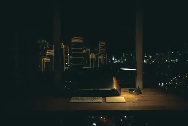 Biurko z laptopem, klawiaturą i lampką nocną. Za oknem nocne miasto, idealne do pracy w godzinach nocnych.