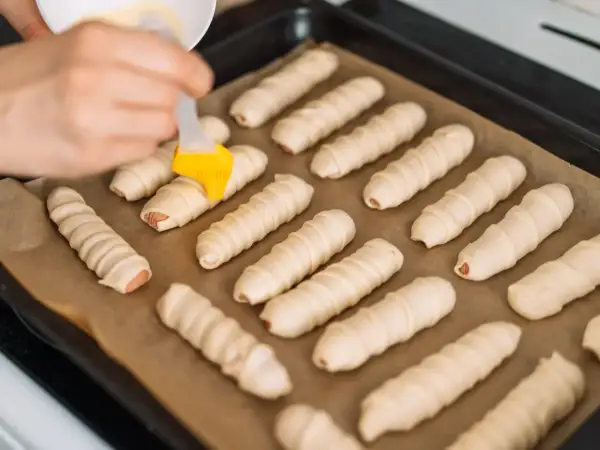 Parówki zawinięte w ciasto francuskie na blasze, przygotowywane do pieczenia.