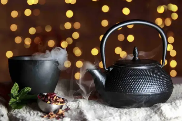 Foto still life: una teiera di ghisa nera emana vapore, accanto a una ciotola fumante e a ingredienti per il tè su uno sfondo di luci soffuse.