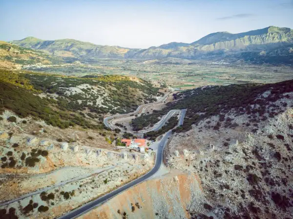 Vue aérienne d'une route sinueuse traversant un paysage montagneux verdoyant, menant à un village sur le plateau de Lassithi. Des ruines de moulins à vent parsèment la colline.