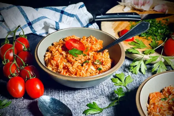 Schüssel mit aromatischem griechischem Reis, garniert mit Tomaten und Petersilie. Frische Tomaten und Knoblauch im Hintergrund.