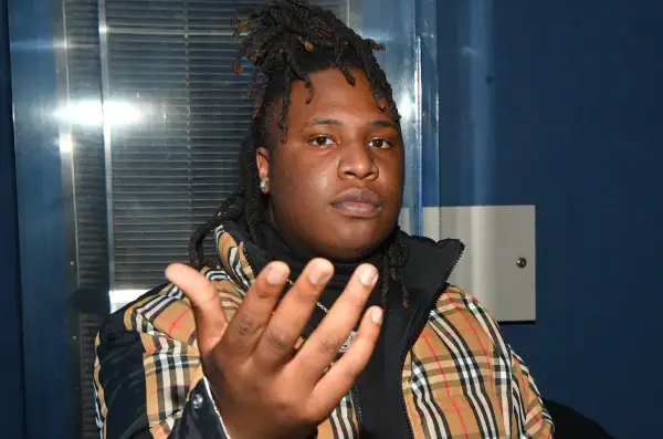 A young man with dreadlocks, wearing a plaid jacket, gestures forward. He's the artist behind many popular Tay Keith songs.
