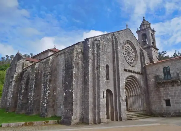 El monasterio de Armenteira, con su imponente fachada de piedra y su rosetón, se alza bajo un cielo azul con nubes.