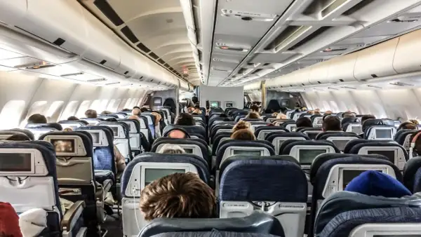 Blick in den Gang eines Flugzeugs. Viele Passagiere sitzen auf ihren Plätzen im Flugzeug innen, einige schauen auf die Bildschirme vor sich.