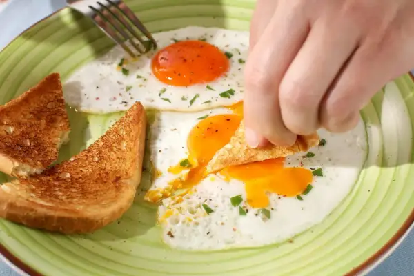 Une main trempe un toast dans un œuf au plat. Combien d'oeuf par jour pour un tel petit-déjeuner ?