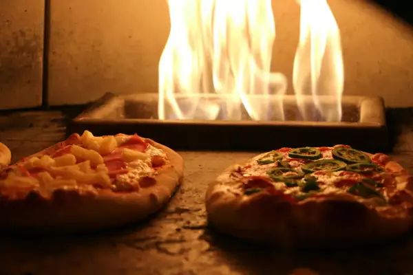 Deux pizzas maison, une à l'ananas et jambon, l'autre aux poivrons verts, cuisent dans un four avec des flammes vives. Une recette pizza maison avec pâte toute prête facile et rapide.