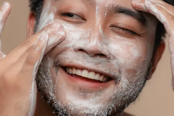 Un visage d'homme souriant, couvert de mousse nettoyante, se fait un soin du visage.