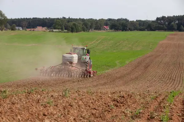 Traktor z siewnikiem HORSCH rozsiewa nawóz, prawdopodobnie fosforan, na polu. W tle las i domy.