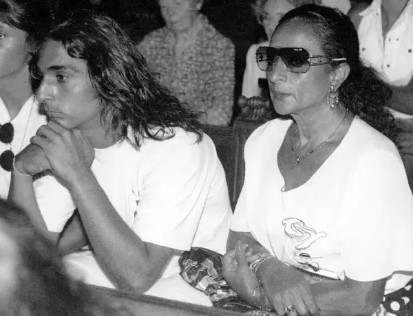 Un joven con pelo largo y una mujer con gafas de sol y joyas, sentados juntos. La imagen evoca la tristeza y el misterio en torno a como murio Lola Flores.