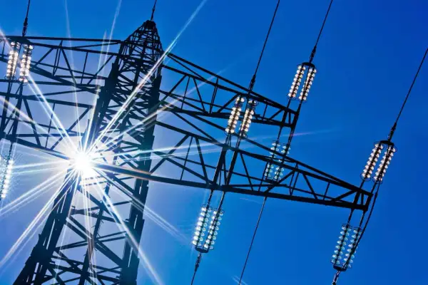 Słońce przebija się przez kratownicę wieży energetycznej, oświetlając izolatory przewodów, które przenoszą prąd dwufazowy.