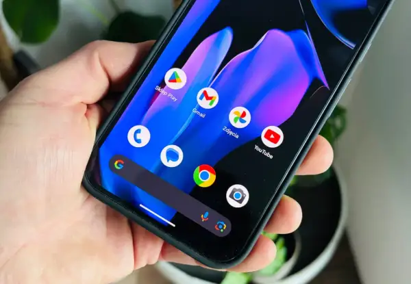 Dłoń trzyma smartfon z kolorową tapetą i ikonami aplikacji, w tym Sklep Play, Gmail, Zdjęcia i YouTube. Wygląda jak nowy android 15.