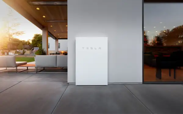 Un elegante Tesla Powerwall blanco se alza junto a una casa moderna con un patio y muebles de exterior.