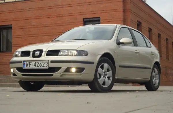 Jasny Seat Leon 1 z włączonymi światłami mijania i przeciwmgielnymi, zaparkowany przed ceglanym budynkiem.