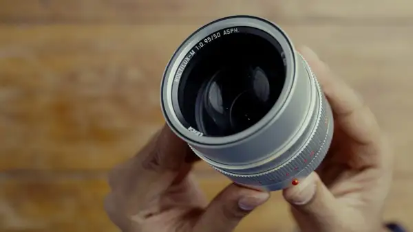 Hands hold a Leica Noctilux M 50 mm lens, its silver barrel and prominent f/0.95 aperture gleaming.