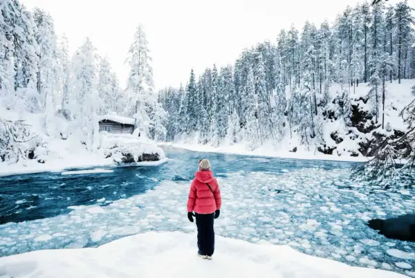 Une personne en manteau rouge admire un paysage hivernal de la pays Lapone, avec une rivière gelée et une cabane enneigée.