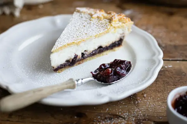 Una fetta di crostata ricotta e visciole, cosparsa di zucchero a velo, su un piatto bianco. Un cucchiaino è pieno di visciole sciroppate.