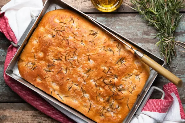 En gyllene focaccia rosmarin i en plåt, toppad med örter och flingsalt. En kniv ligger på brödet, redo att skäras.