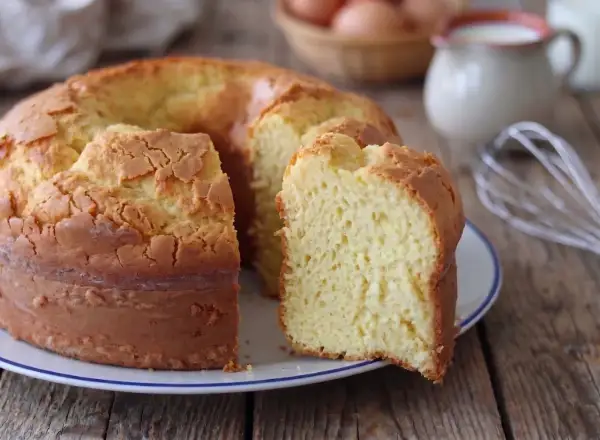 Una torta alta, soffice e dorata, con una fetta tagliata che rivela la sua morbidezza. Sullo sfondo, uova e latte, ingredienti per un dolce fatto in casa.