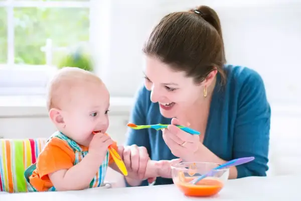 Mama karmi niemowlę, które próbuje samodzielnie jeść. To obrazek z tabeli żywienia 7-miesięcznego dziecka.