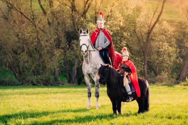 Zwei Reiter in römischer Kleidung, ein Mann auf einem Schimmel und ein Kind auf einem Pony, reiten im Sonnenschein. Ein Bild wie aus einer St. Martin-Geschichte.