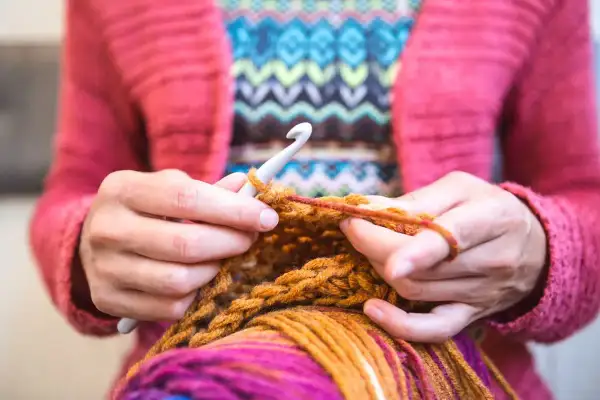 Mani che lavorano a maglia con un uncinetto, filato colorato e un maglione rosa. Nonostante sembri rilassante, lavorare a maglia fa male al cuore se si pensa troppo.