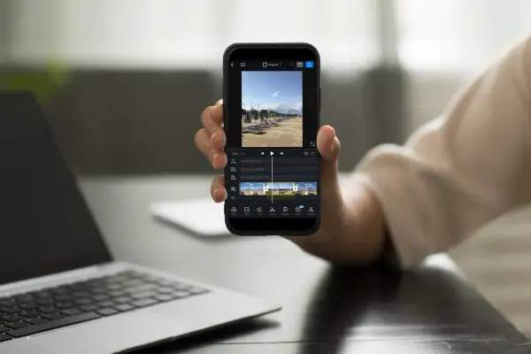 Dłoń trzyma smartfon z aplikacją do montowania filmów. Na ekranie widać oś czasu z klipami wideo i opcjami edycji.