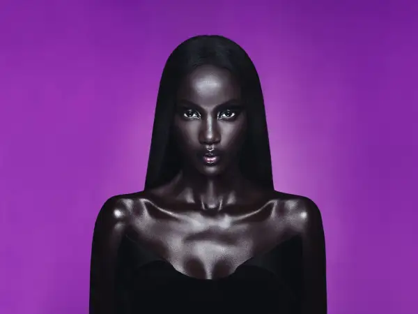 Anok Yai, con pelle lucida e capelli neri, posa con uno sguardo intenso su sfondo viola.