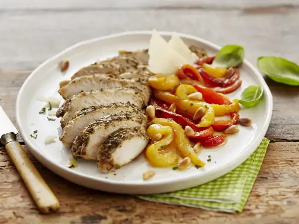 Sabroso pollo con pimientos asados, piñones y queso parmesano, servido en un plato blanco sobre una mesa de madera.