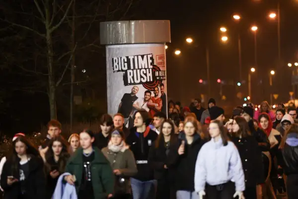 Tłum młodych ludzi zmierza na koncert Big Time Rush w Polsce. Plakat zespołu zdobi słup ogłoszeniowy.