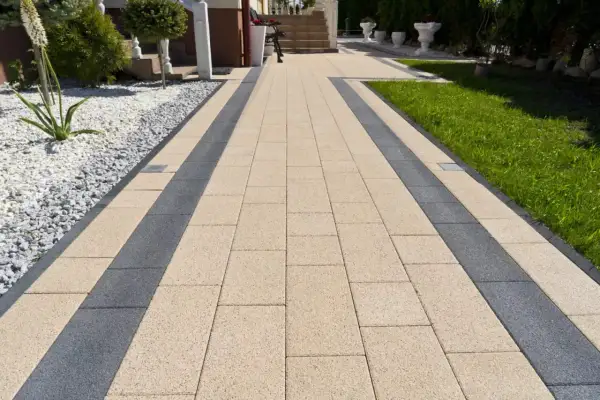 Eleganckie ścieżki z kostki brukowej w beżowo-grafitowym wzorze prowadzą do domu. Otoczone zielenią i białym żwirem.