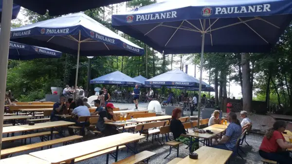 Ein sonniger Tag im Paulaner Biergarten. Gäste sitzen an Holztischen unter großen Sonnenschirmen und genießen die Atmosphäre.