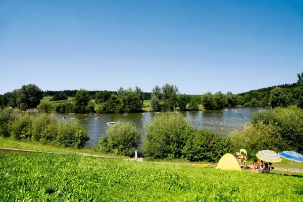 Sonniger Tag am Aichstruter Stausee. Menschen planschen im Wasser, baden und entspannen sich am Ufer unter bunten Sonnenschirmen.