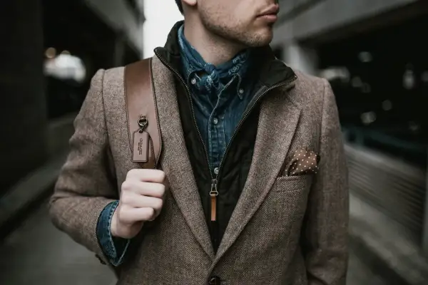 Mann im Tweed-Sakko mit Jeanshemd und Halstuch, perfekt für den party chic dress code male. Braune Ledertasche mit Coach-Anhänger.